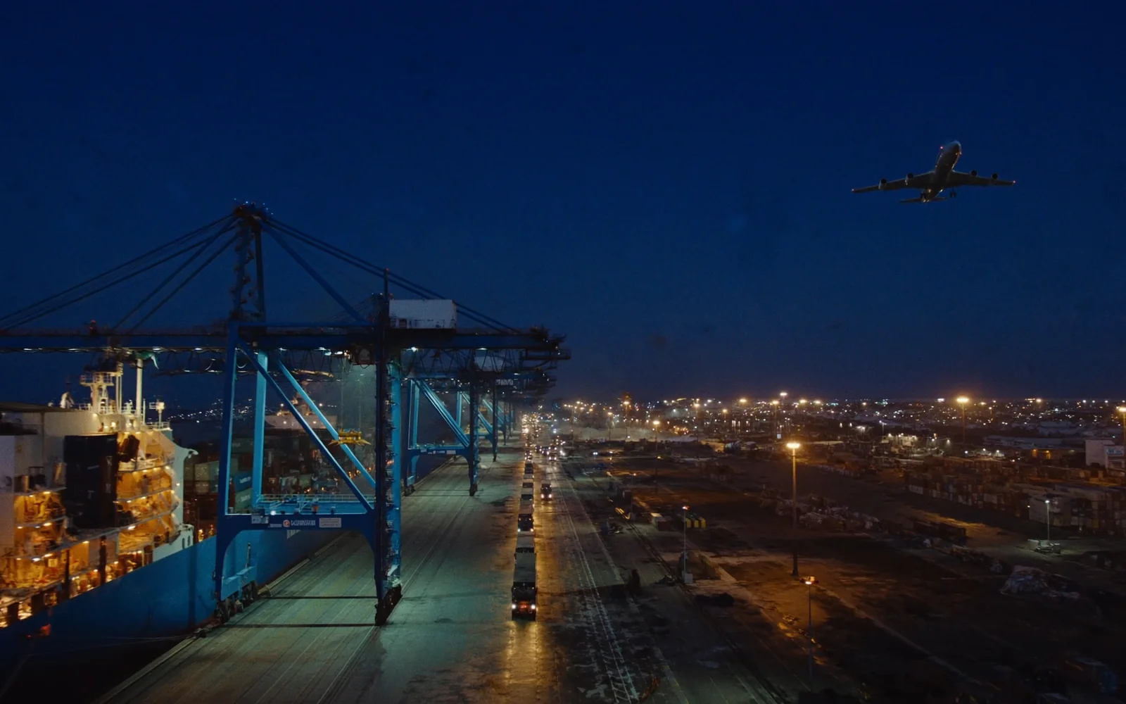 A container terminal at blue hour — a vessel under gantry cranes, a lane of trucks leaving the quay, and a cargo aircraft climbing away